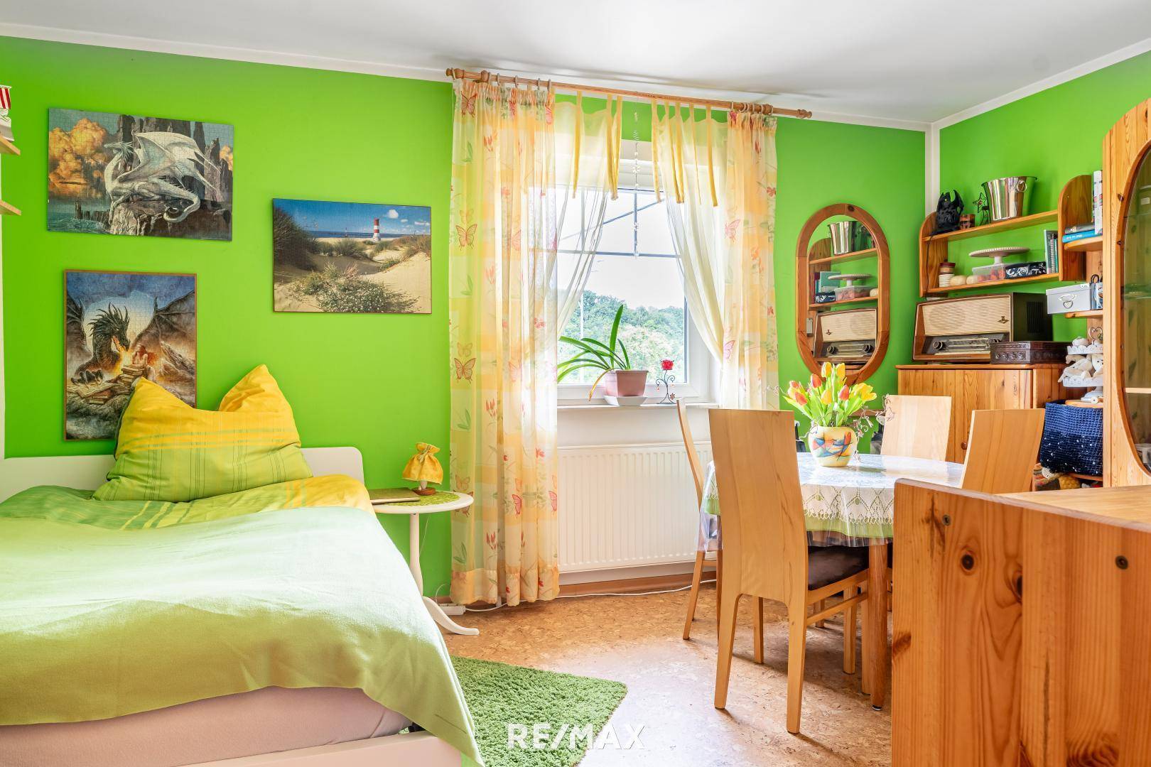 Helles Zimmer mit grünen Wänden, Bett, kleinem Esstisch und Blick ins Grüne durch das Fenster.