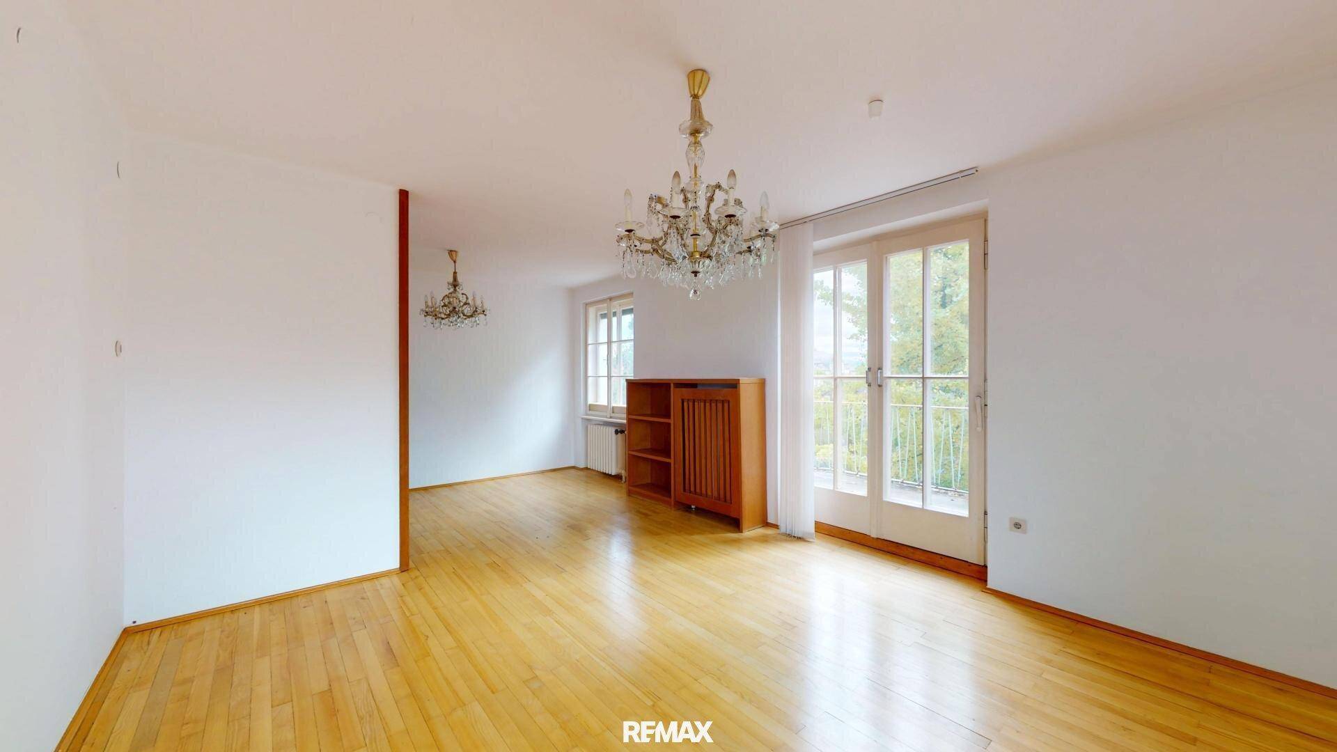 Geräumiges, leeres Zimmer mit Holzboden, Kronleuchter und Zugang zum Balkon.