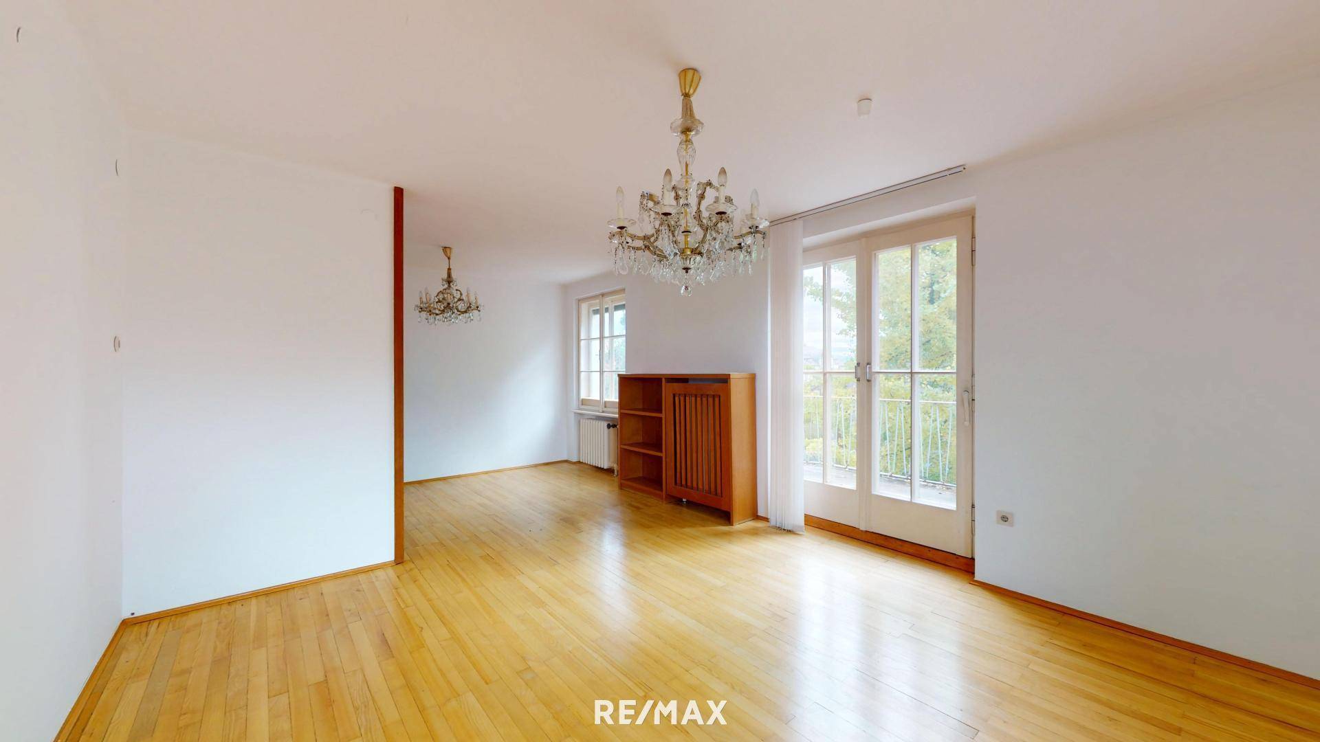 Leeres Zimmer mit Parkettboden, Kronleuchter und Zugang zu einem französischen Balkon.