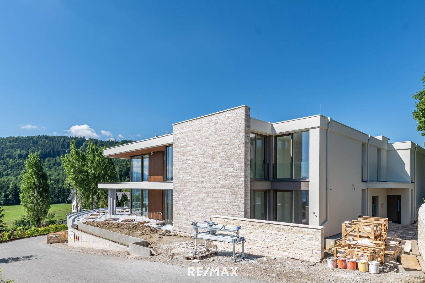 Modernes Gebäude in Bauphase mit Steinfassade und großen Fensterfronten vor grüner Hügellandschaft.
