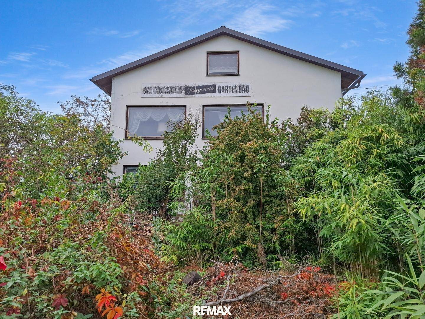 Rückansicht des Hauses mit üppiger Vegetation und einem Schild für Gartenbau unter blauem Himmel.