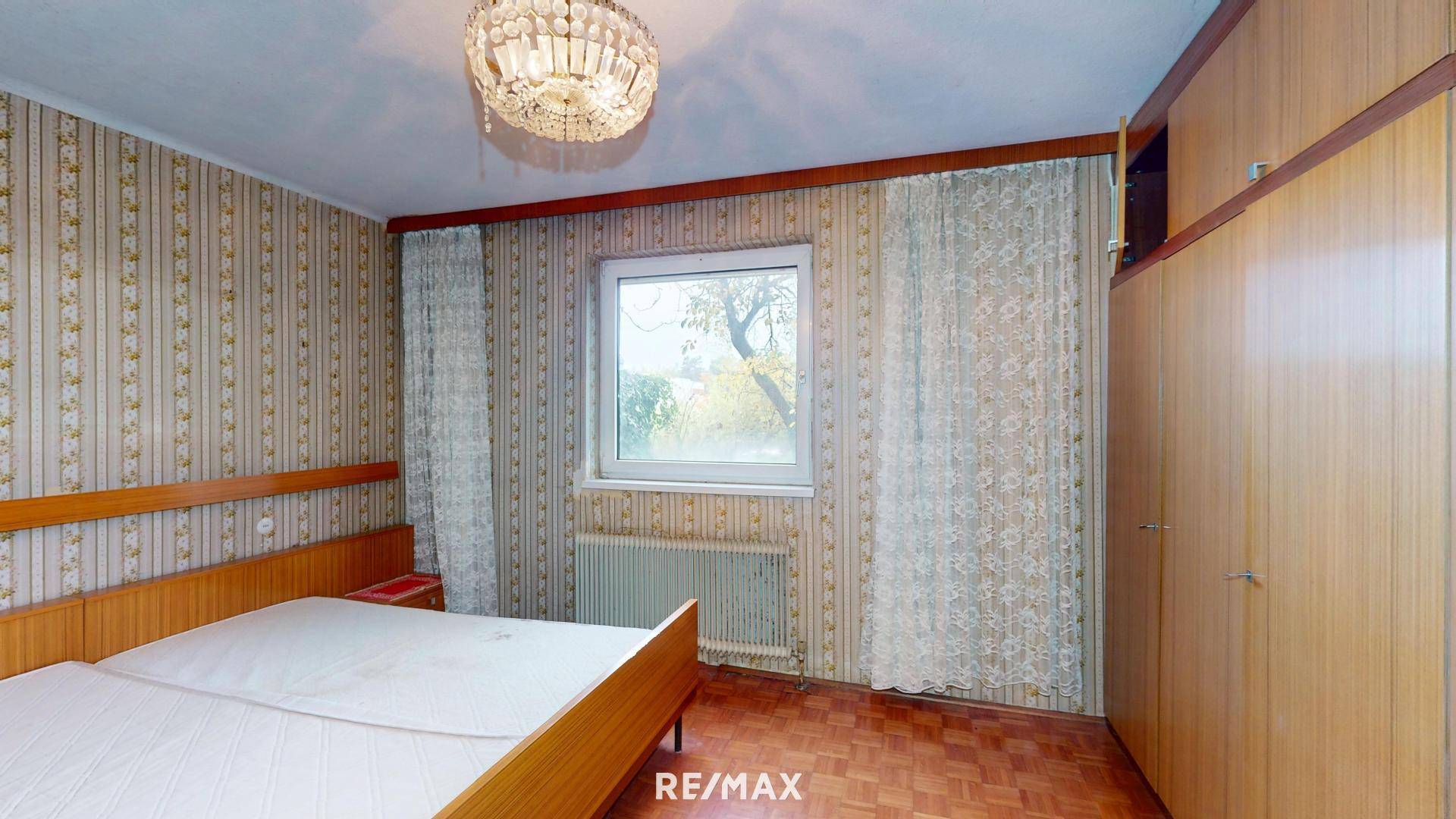 Schlafzimmer mit Doppelbett, Holzschrank und gemusterter Tapete, hell durch ein großes Fenster.