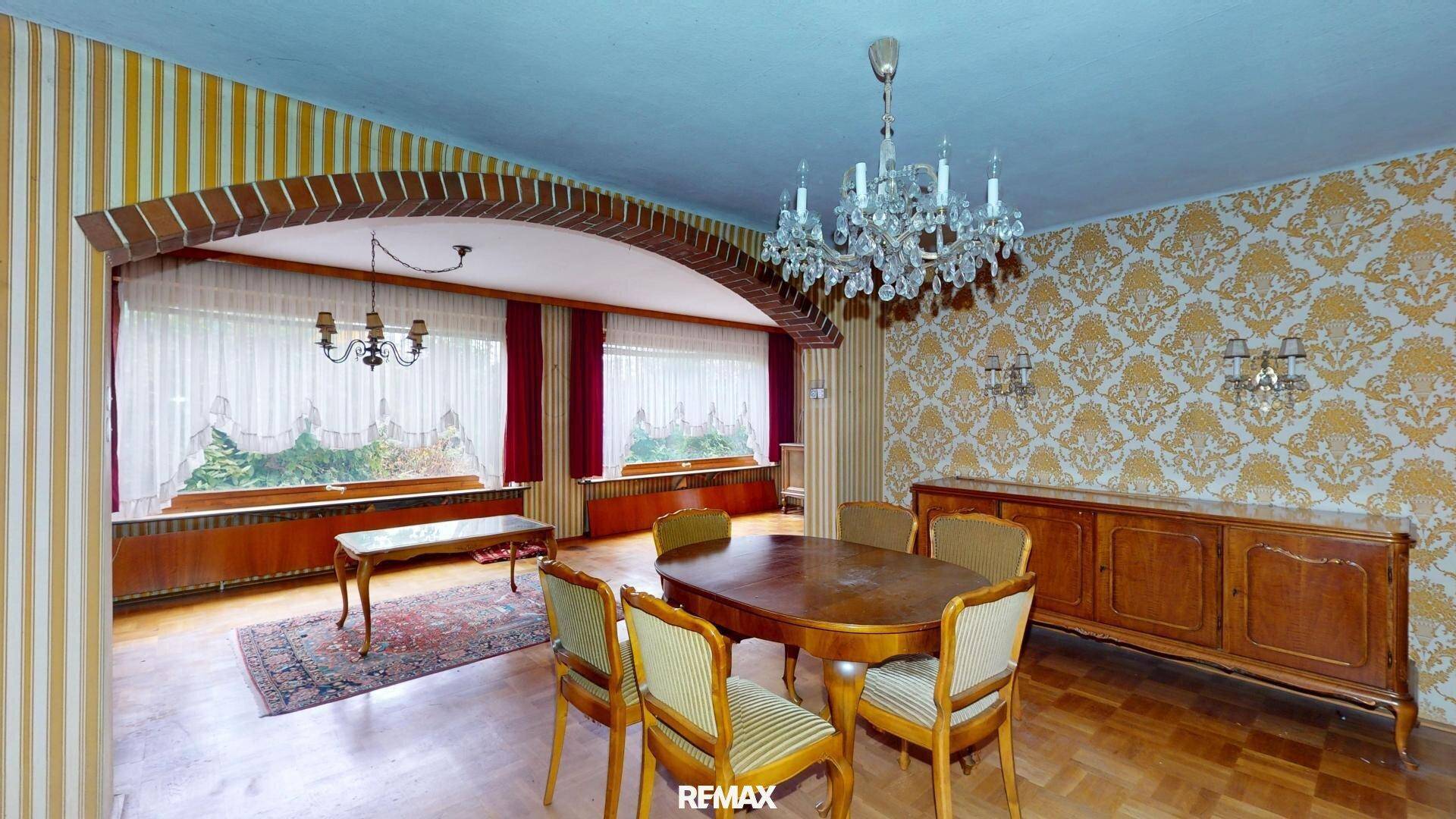 Klassisches Esszimmer mit Kronleuchter, Holztisch und gemusterten Tapeten, durch einen Bogen getrennt.