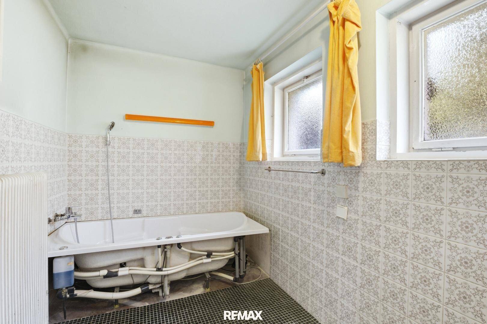 Badezimmer mit Badewanne, gefliesten Wänden und zwei Fenstern, sanierungsbedürftig.