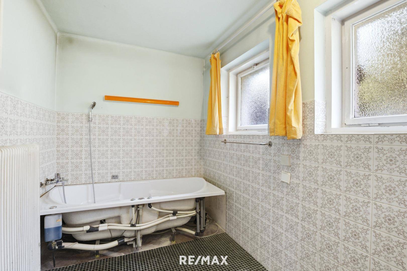 Badezimmer mit Badewanne, Fliesen an den Wänden und zwei Fenstern.