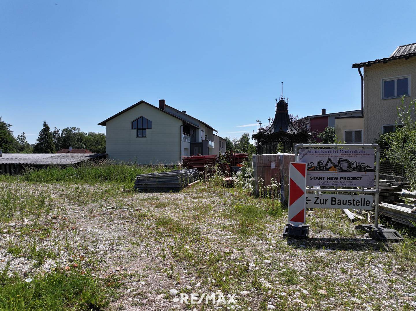 Unbebautes Grundstück mit Baustellenschild und angrenzenden Häusern unter klarem Himmel.