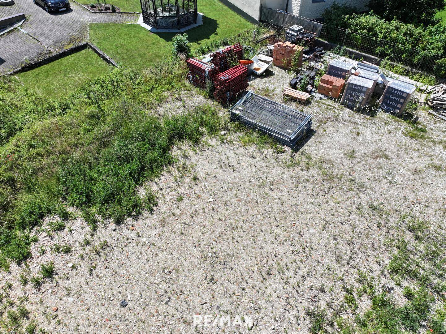 Luftaufnahme eines unbebauten Grundstücks mit Baumaterialien, angrenzend an einen Garten mit Pavillon.