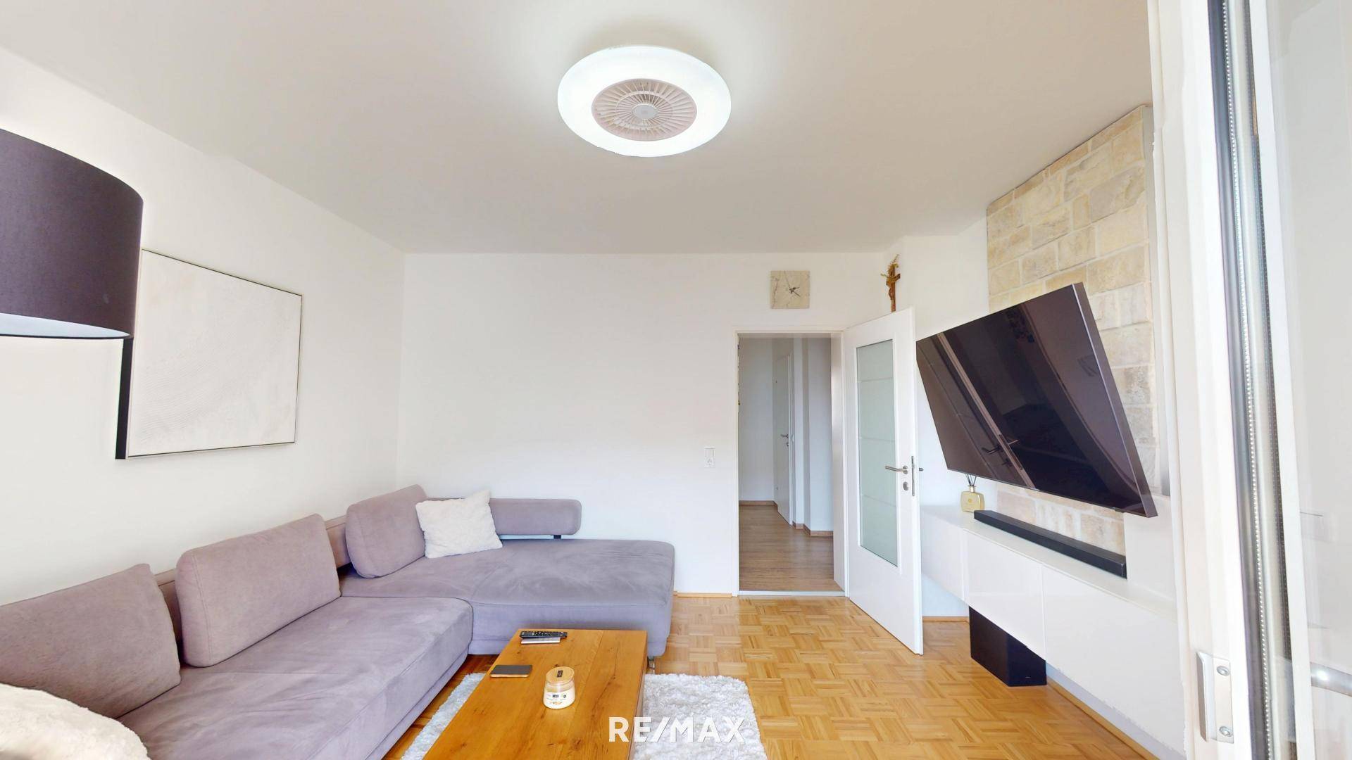 Wohnzimmer mit Fernseher, Parkettboden und Zugang zu einem Balkon oder Terrasse.