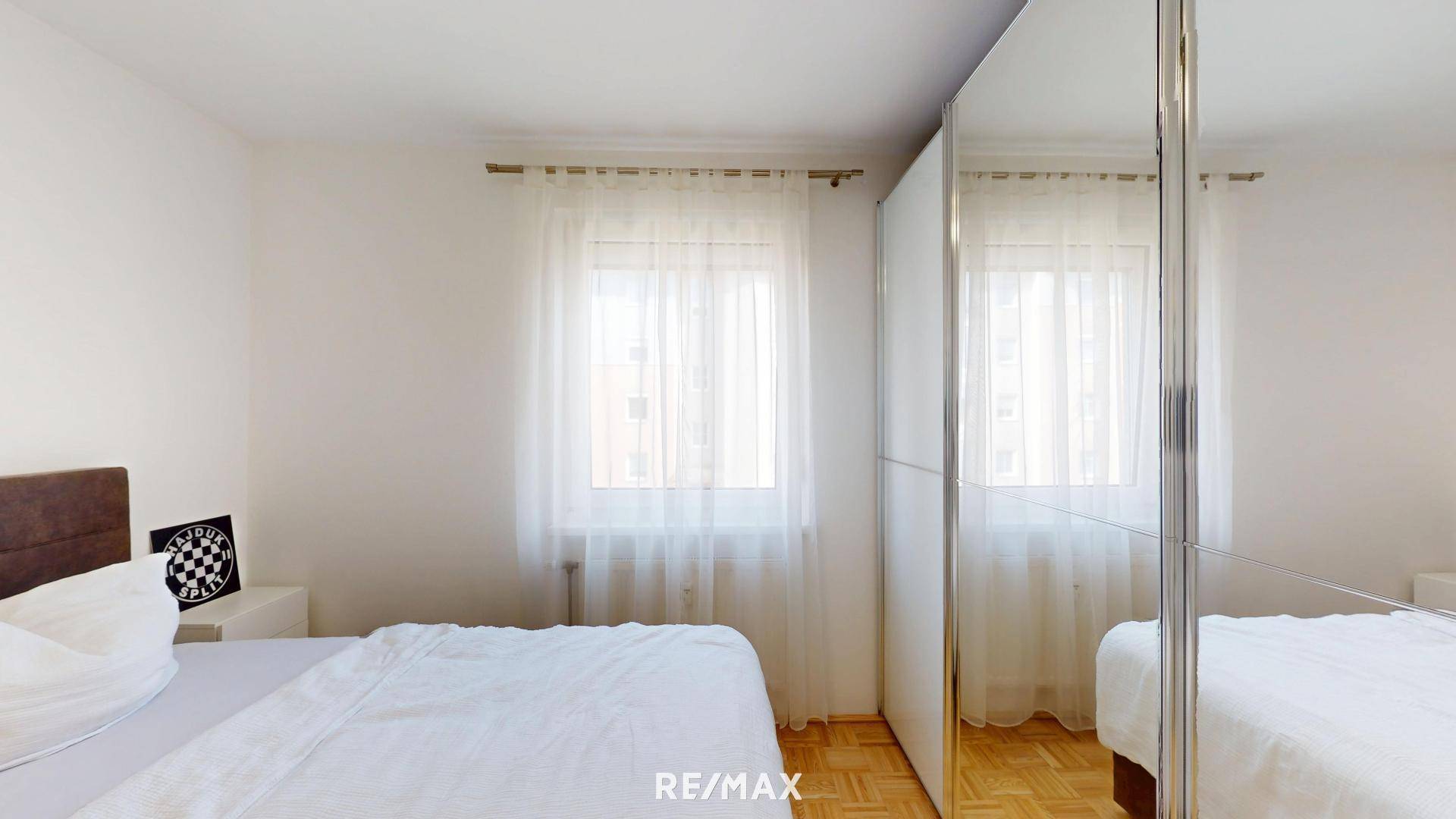 Schlafzimmer mit Parkettboden, Fenster und Spiegelschrank.