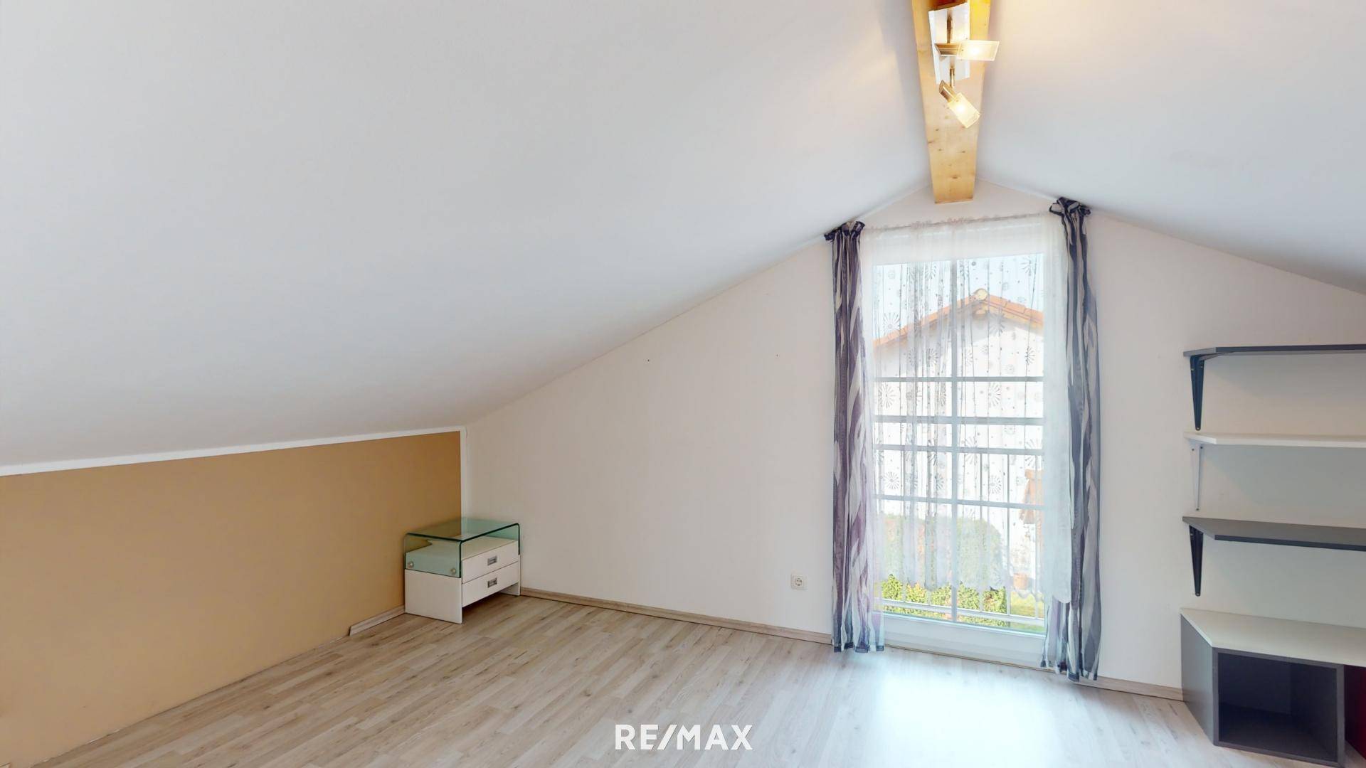 Dachgeschosszimmer mit schrägen Wänden, Holzbalken und Laminatboden, hell durch großes Fenster.