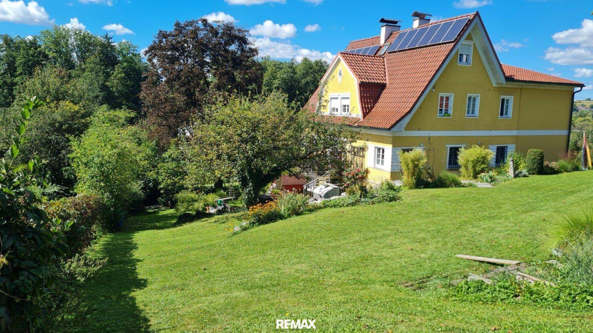 Weitläufiger Garten mit gelbem Wohnhaus, roten Dachziegeln und Solaranlage unter blauem Himmel.