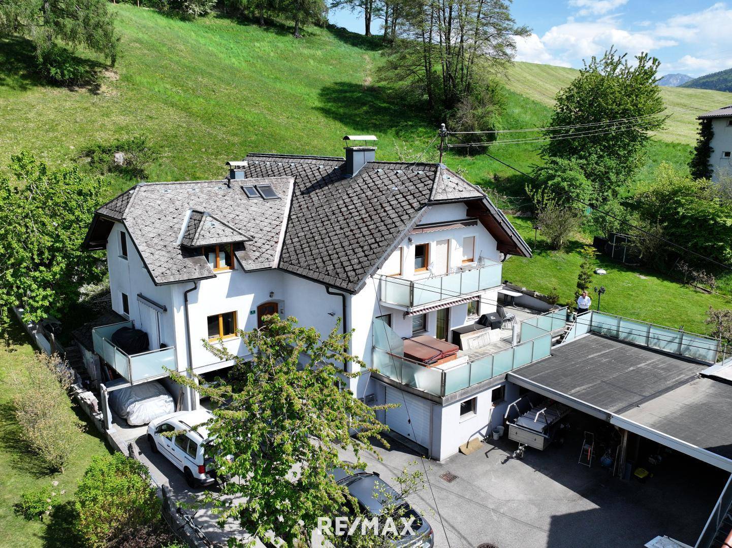 Luftaufnahme des Hauses mit Satteldach, Balkonen, Whirlpool und Carport, umgeben von grünen Hügeln.