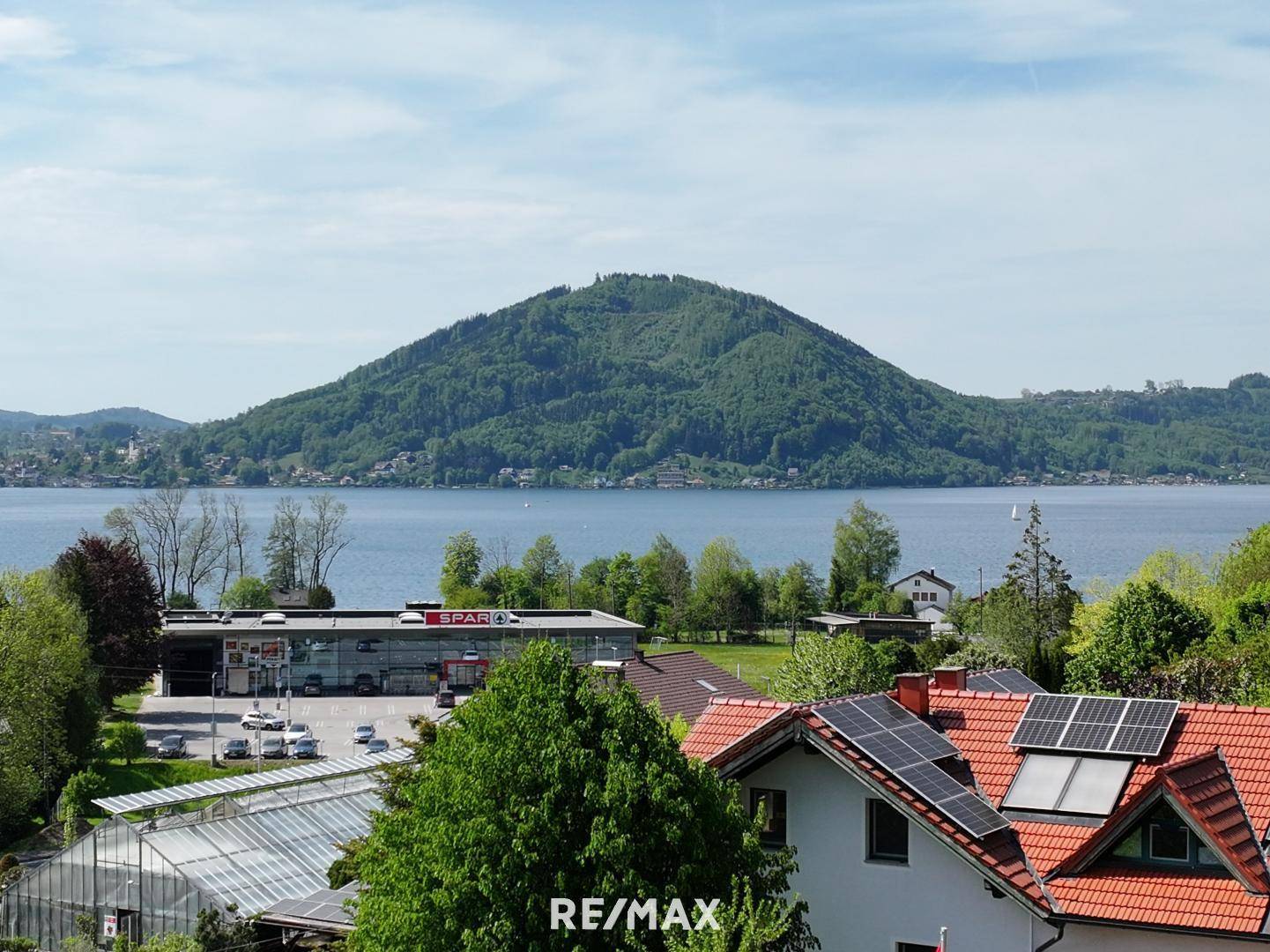 Blick über Dächer mit Solaranlagen auf einen See und eine grüne Berglandschaft.