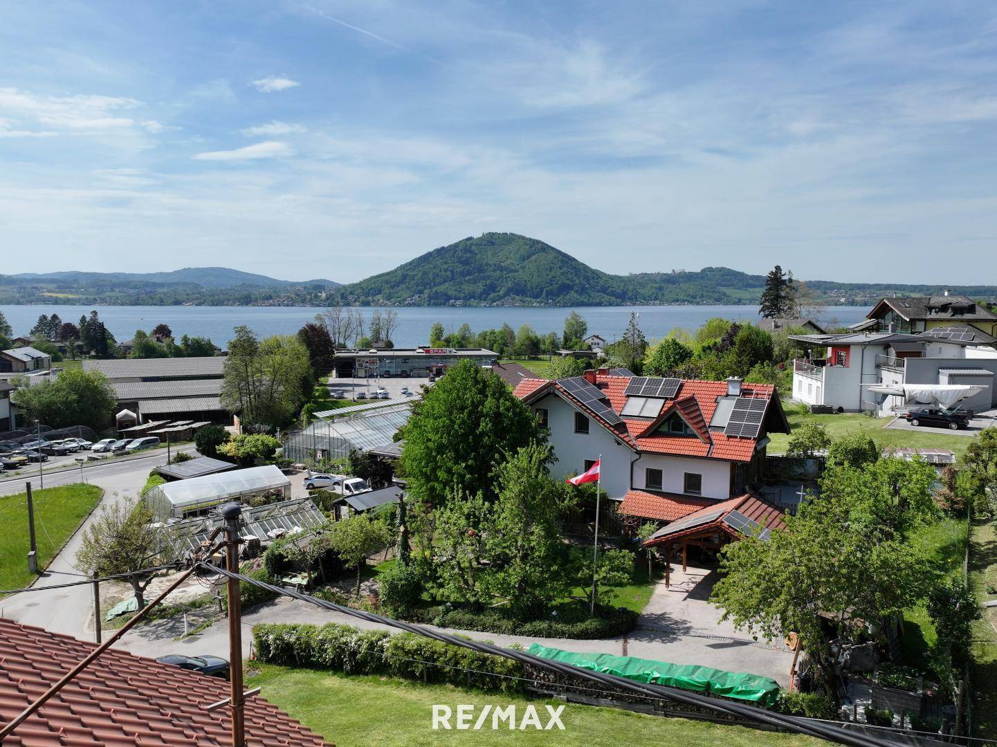Luftaufnahme eines Hauses mit Solaranlagen, umgeben von Grünflächen und einem See mit Bergen.