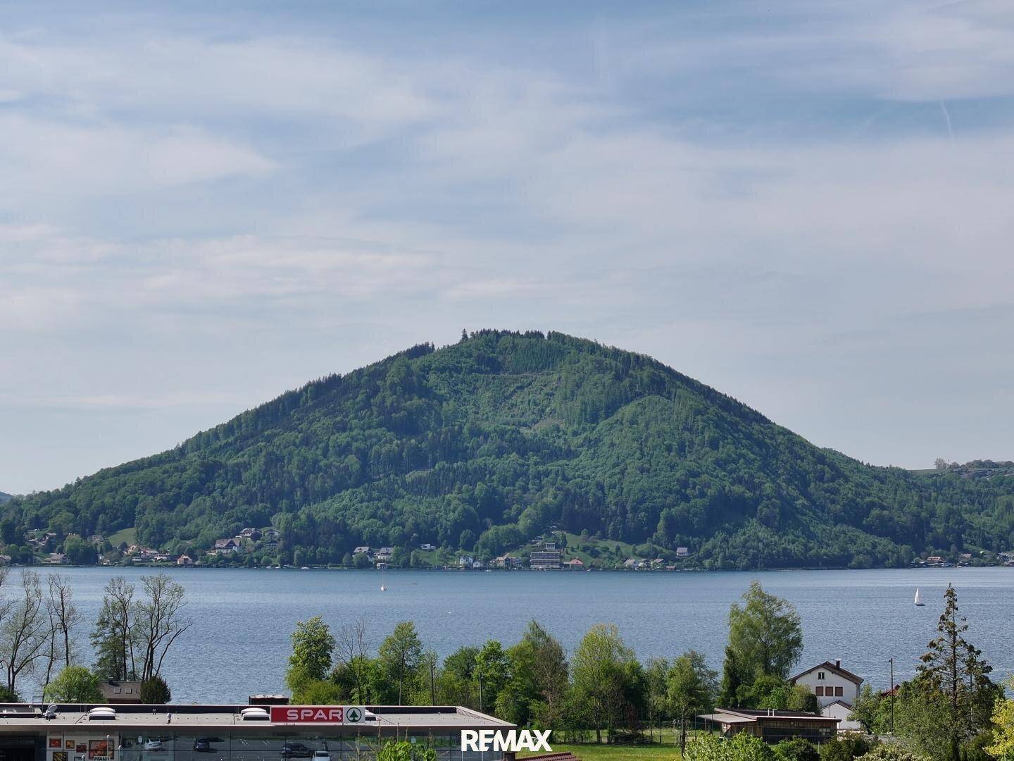 Weitläufiger Blick über den See und die umliegende Landschaft mit einem Supermarkt und Dächern mit Solaranlagen.