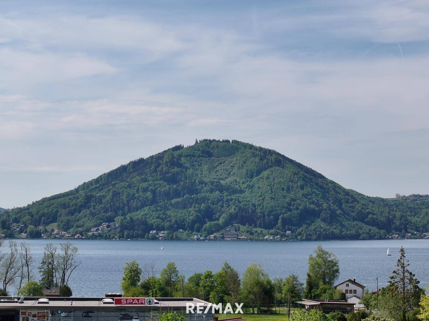 Weitläufige Aussicht auf den See und die bewaldeten Berge unter einem blauen Himmel.