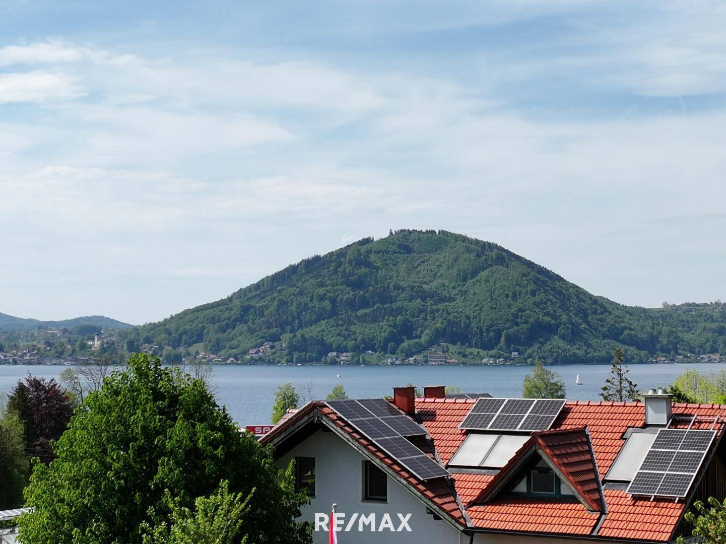 Dächer mit Solarmodulen im Vordergrund und ein malerischer See mit Bergen im Hintergrund.