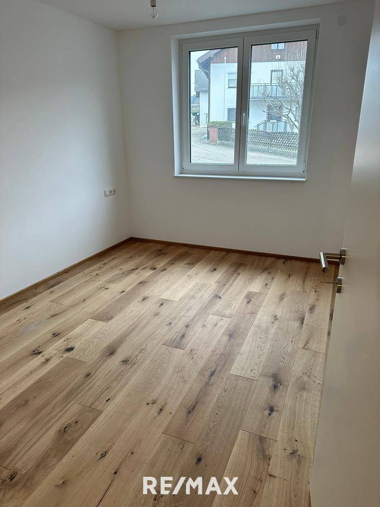 Leeres Zimmer mit Holzboden und großem Fenster, das viel Tageslicht hereinlässt.