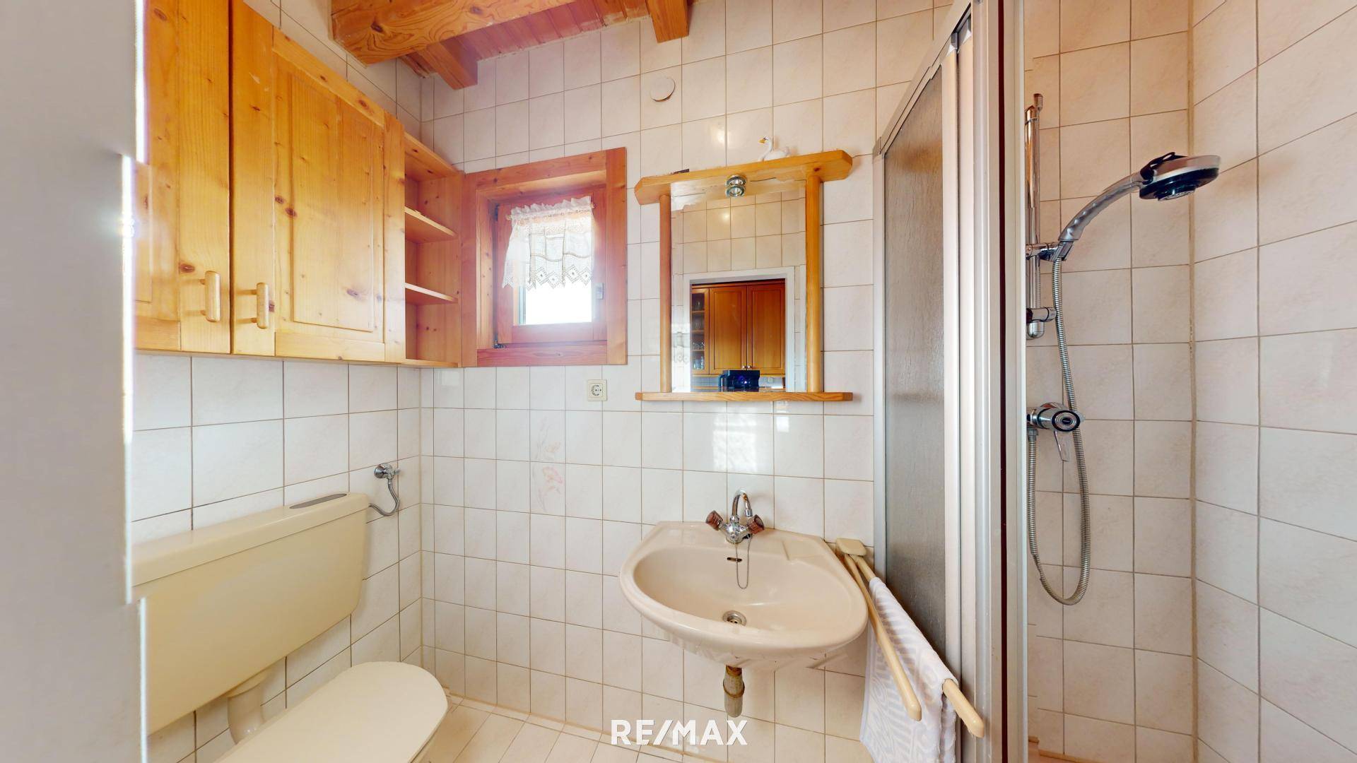 Einfaches Badezimmer mit weißen Fliesen, Dusche, WC, Waschbecken und kleinem Fenster.