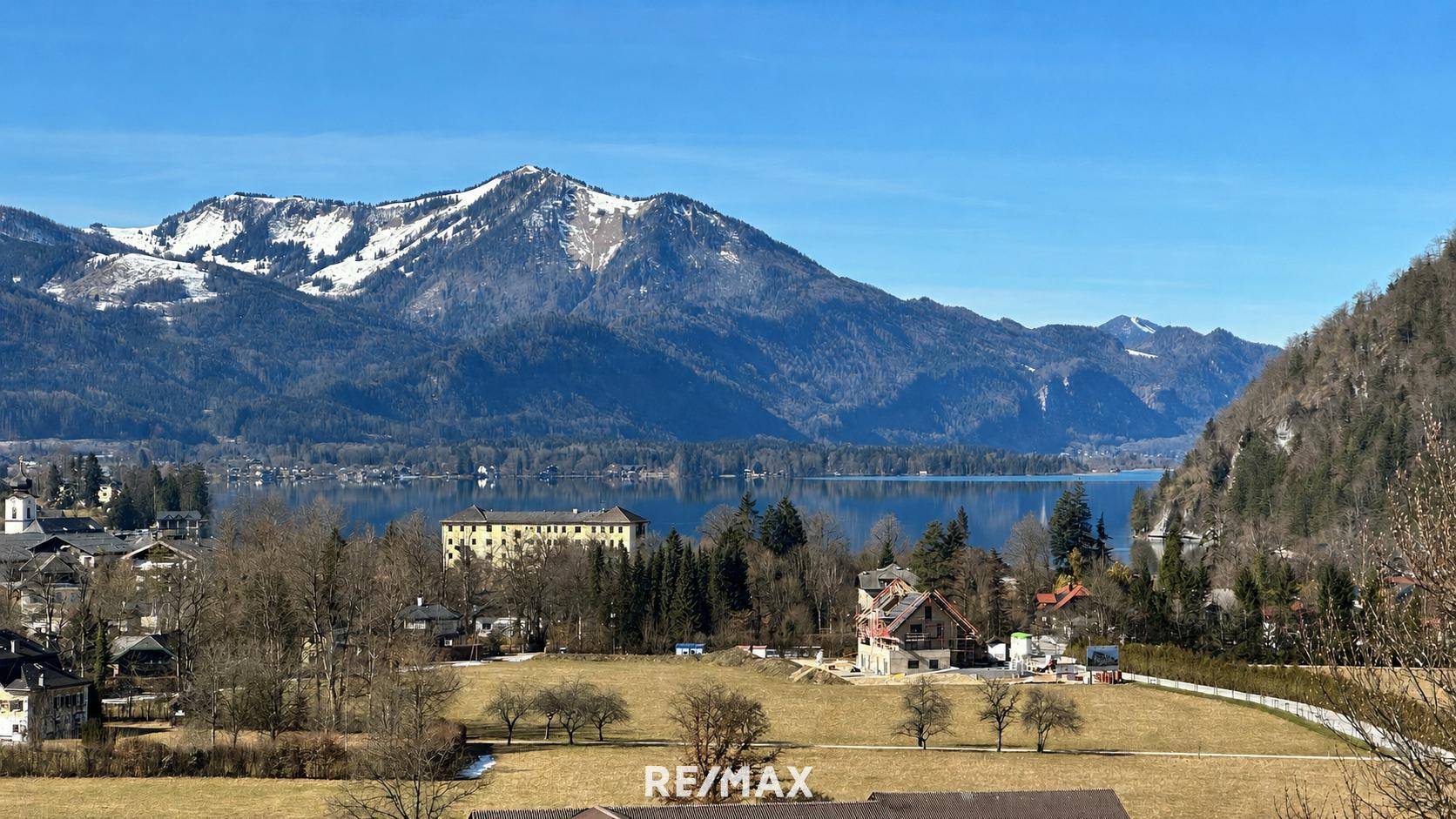 Sonniger Panoramablick auf den Wolfgangsee mit schneebedeckten Bergen und grünen Wiesen.