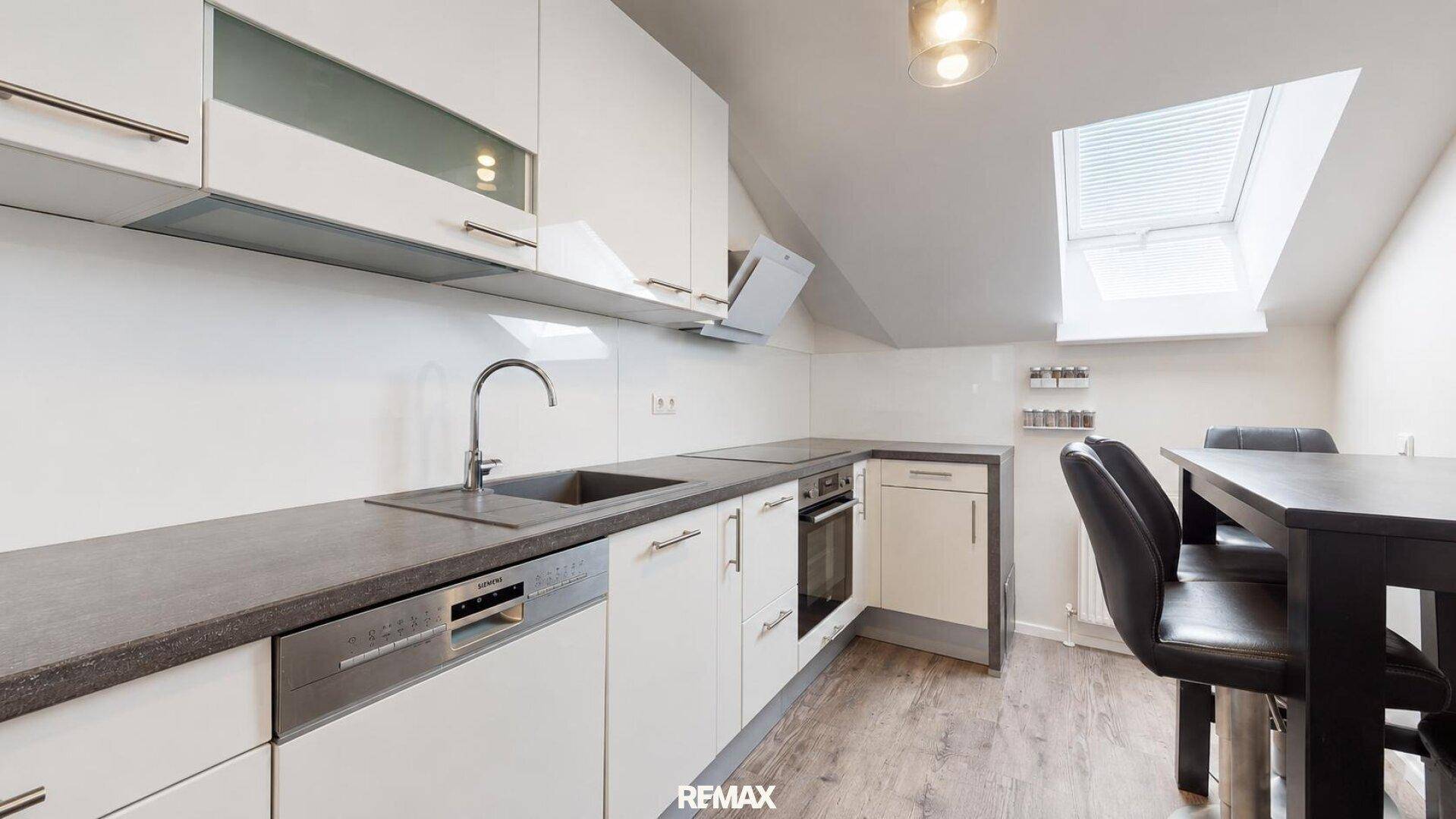 Moderne Einbauküche mit weißen Schränken, dunkler Arbeitsplatte, Essbereich und Dachfenster.