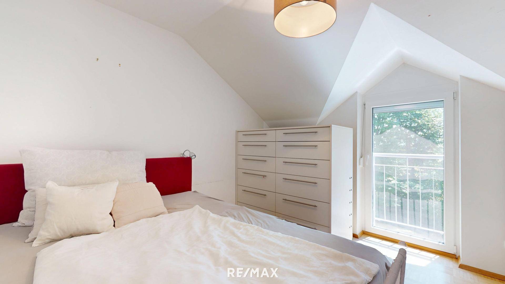 Schlafzimmer mit Bett, Kommode und Zugang zu einem französischen Balkon unter einer Dachschräge.