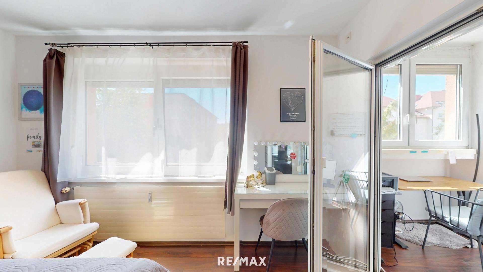 Helles Schlafzimmer mit großem Fenster, Schminktisch und einer Glastrennwand zu einem weiteren Bereich.