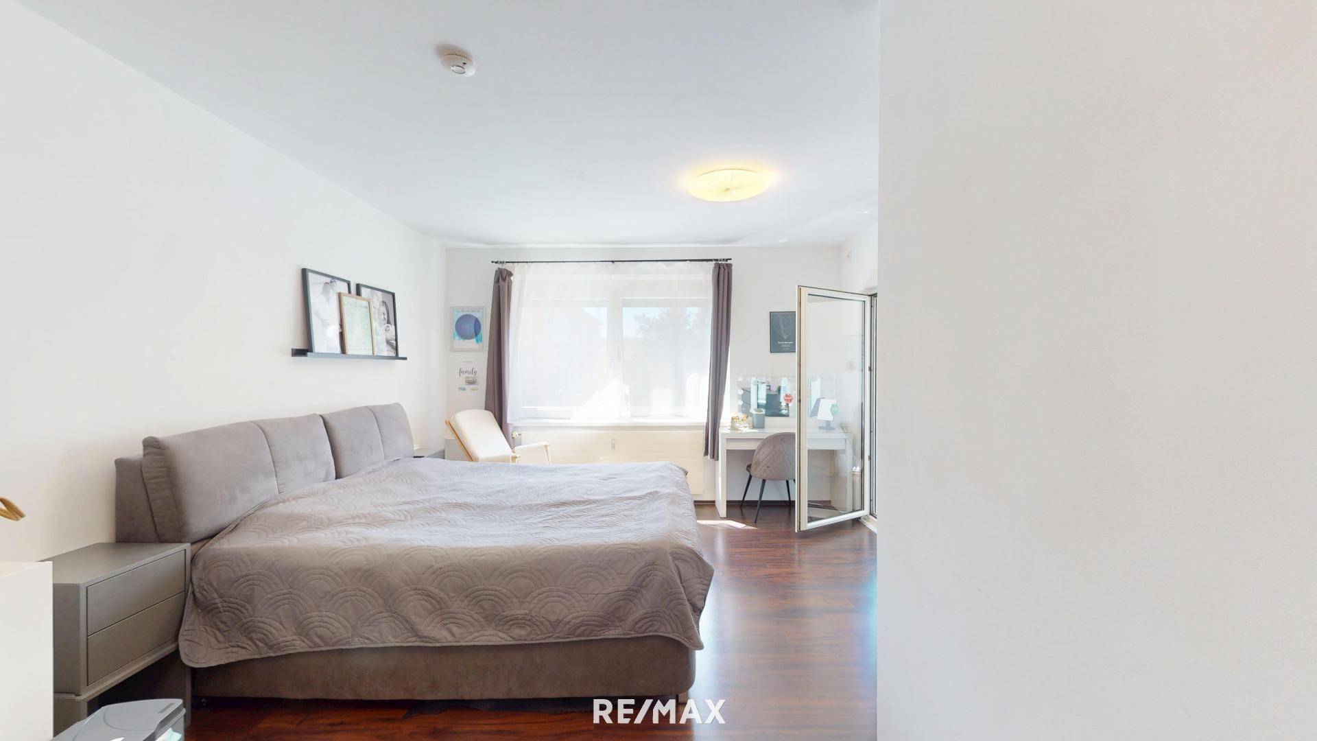 Helles Schlafzimmer mit großem Bett, dunklem Holzboden und einem Arbeitsbereich am Fenster.