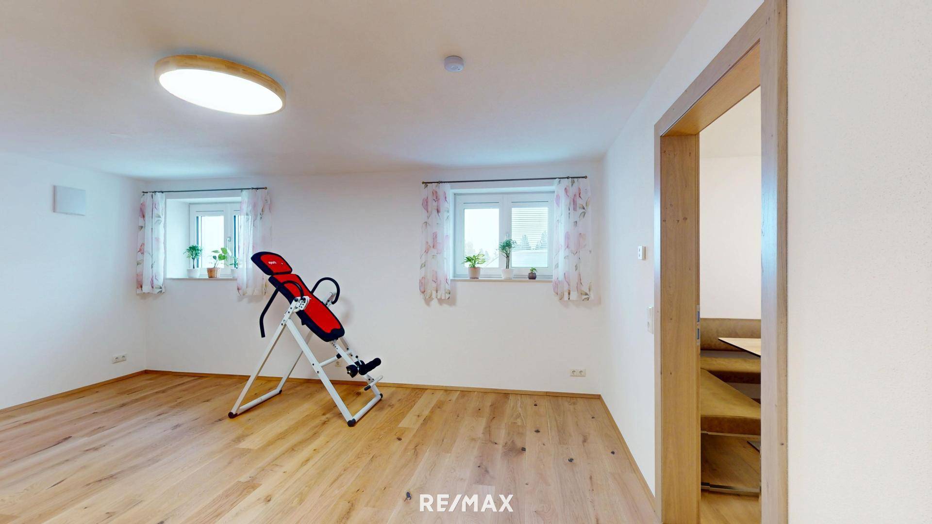 Heller Mehrzweckraum mit Holzboden, zwei Fenstern und einem Trainingsgerät, ideal für Fitnessaktivitäten.