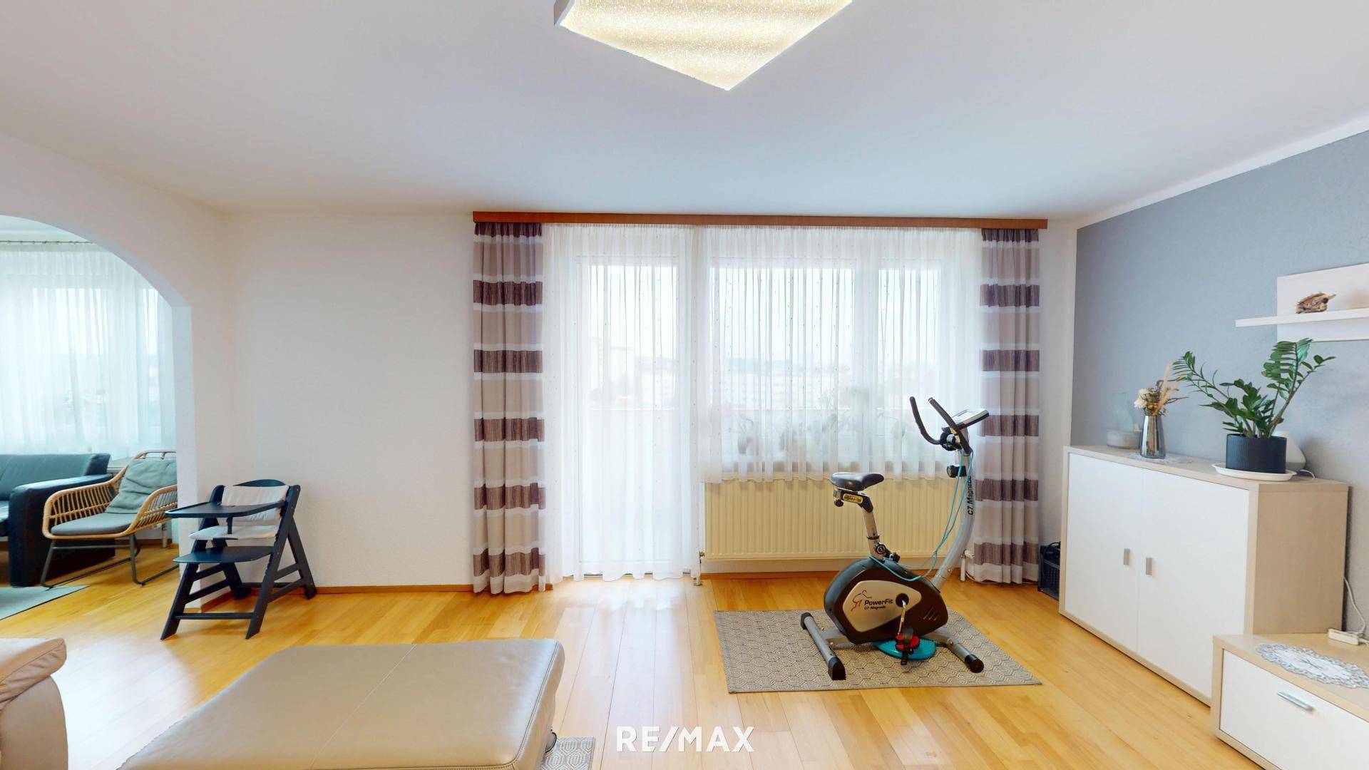 Heller Wohnbereich mit Trainingsfahrrad, großen Fenstern und Holzboden.