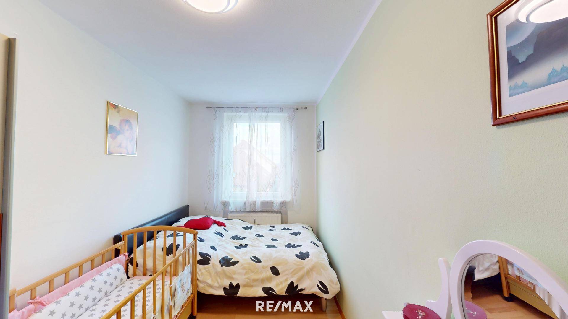 Schlafzimmer mit Doppelbett, Babybett, Schminktisch und Fenster mit hellem Vorhang.