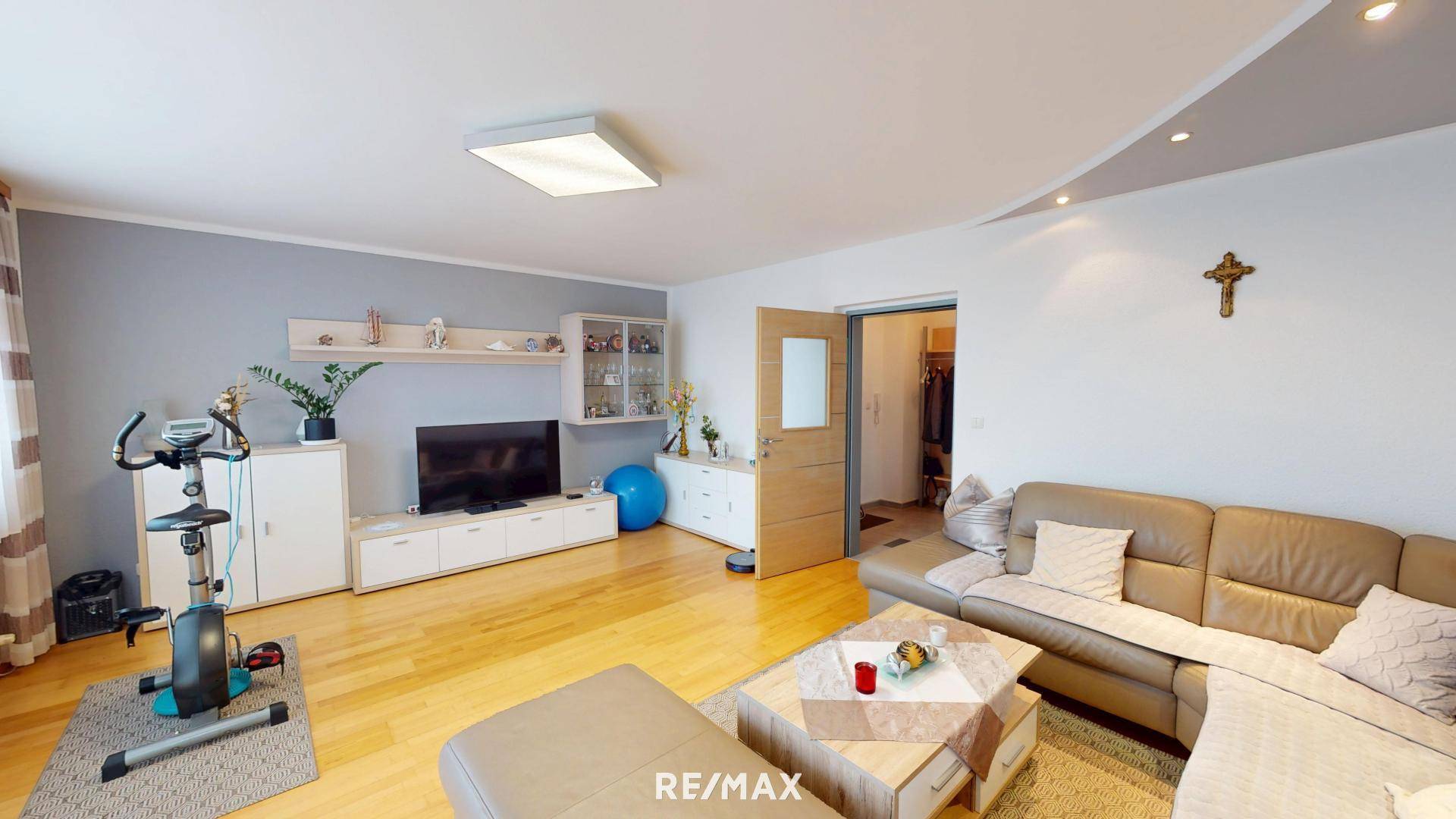 Geräumiges Wohnzimmer mit Ecksofa, Fernseher, Trainingsfahrrad und Holzboden.