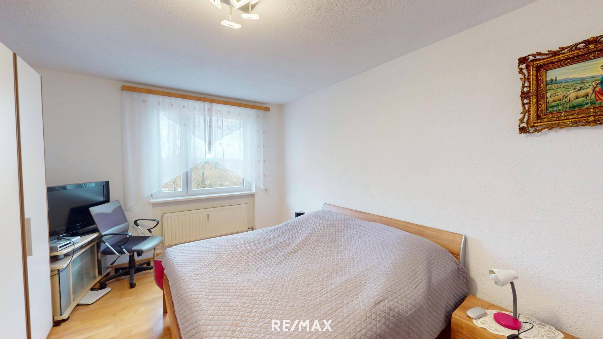 Schlafzimmer mit Doppelbett, Kleiderschrank, Schreibtisch und Fenster mit hellem Vorhang.