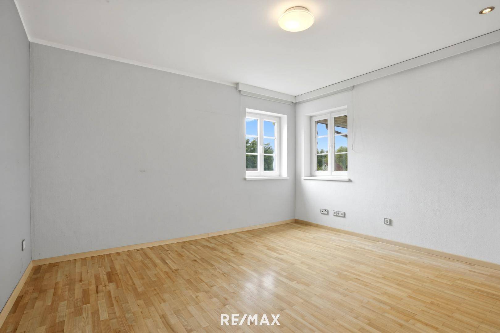 Geräumiges Schlafzimmer mit hellem Holzboden und zwei Fenstern mit Ausblick.