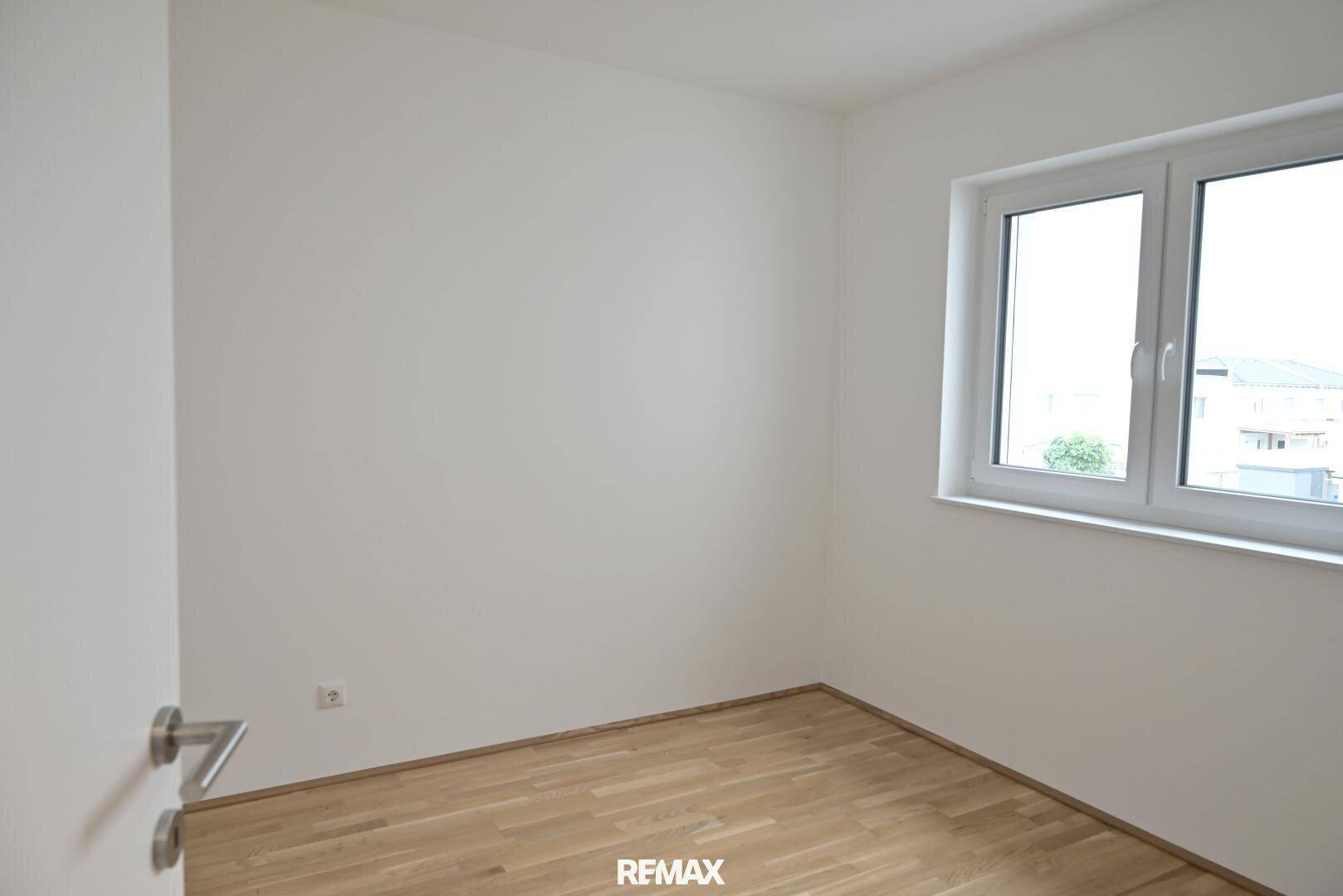 Helles, leeres Schlafzimmer mit Holzboden und einem Fenster, das Tageslicht hereinlässt.