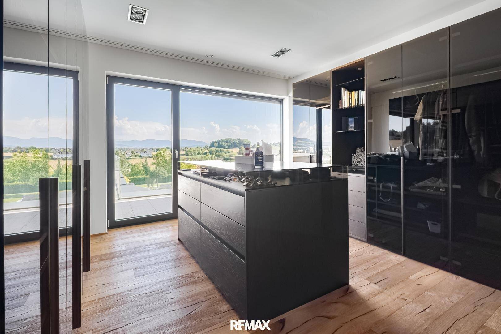 Geräumiger begehbarer Kleiderschrank mit dunklen Einbauschränken, Mittelinsel und großen Fenstern.