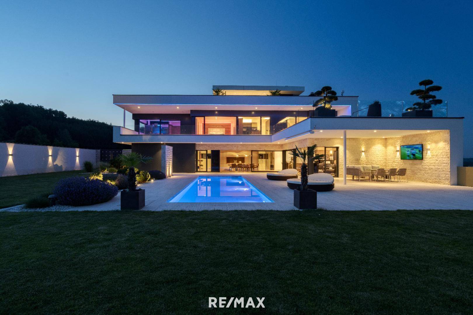 Moderne Luxusvilla bei Dämmerung mit beleuchtetem Infinity-Pool, Terrasse und gepflegtem Garten.