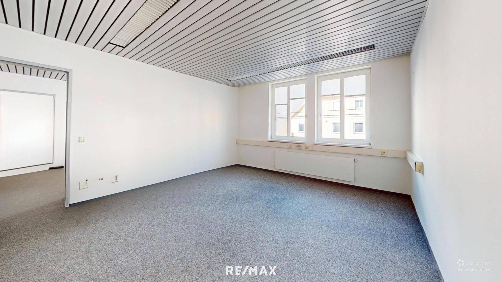 Heller Büroraum mit Teppichboden, Lamellendecke und Fenstern, die Tageslicht hereinlassen.
