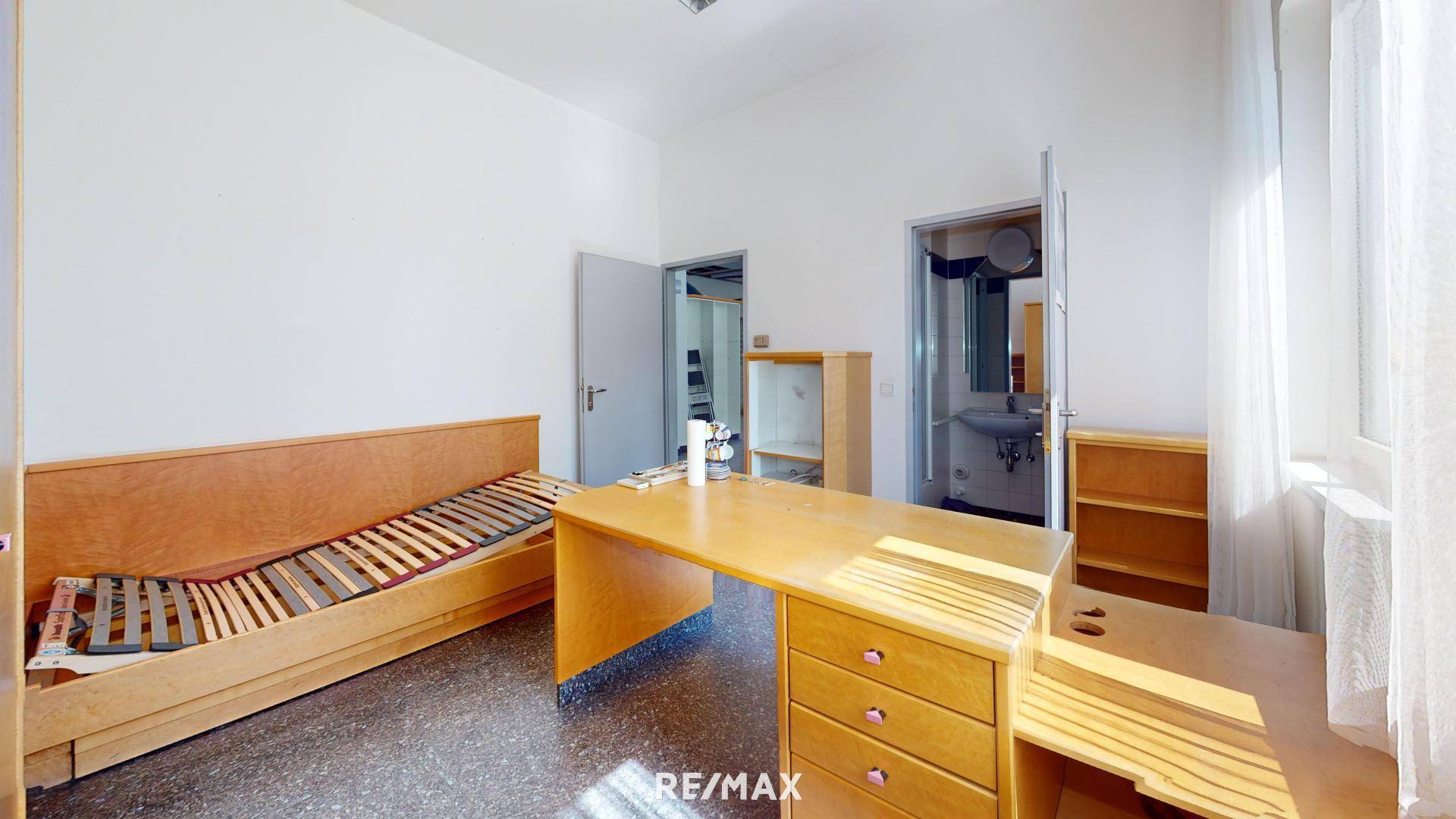 Kleiner Büroraum mit Holzmöbeln, einem Bettgestell und Zugang zu einem Badezimmer.