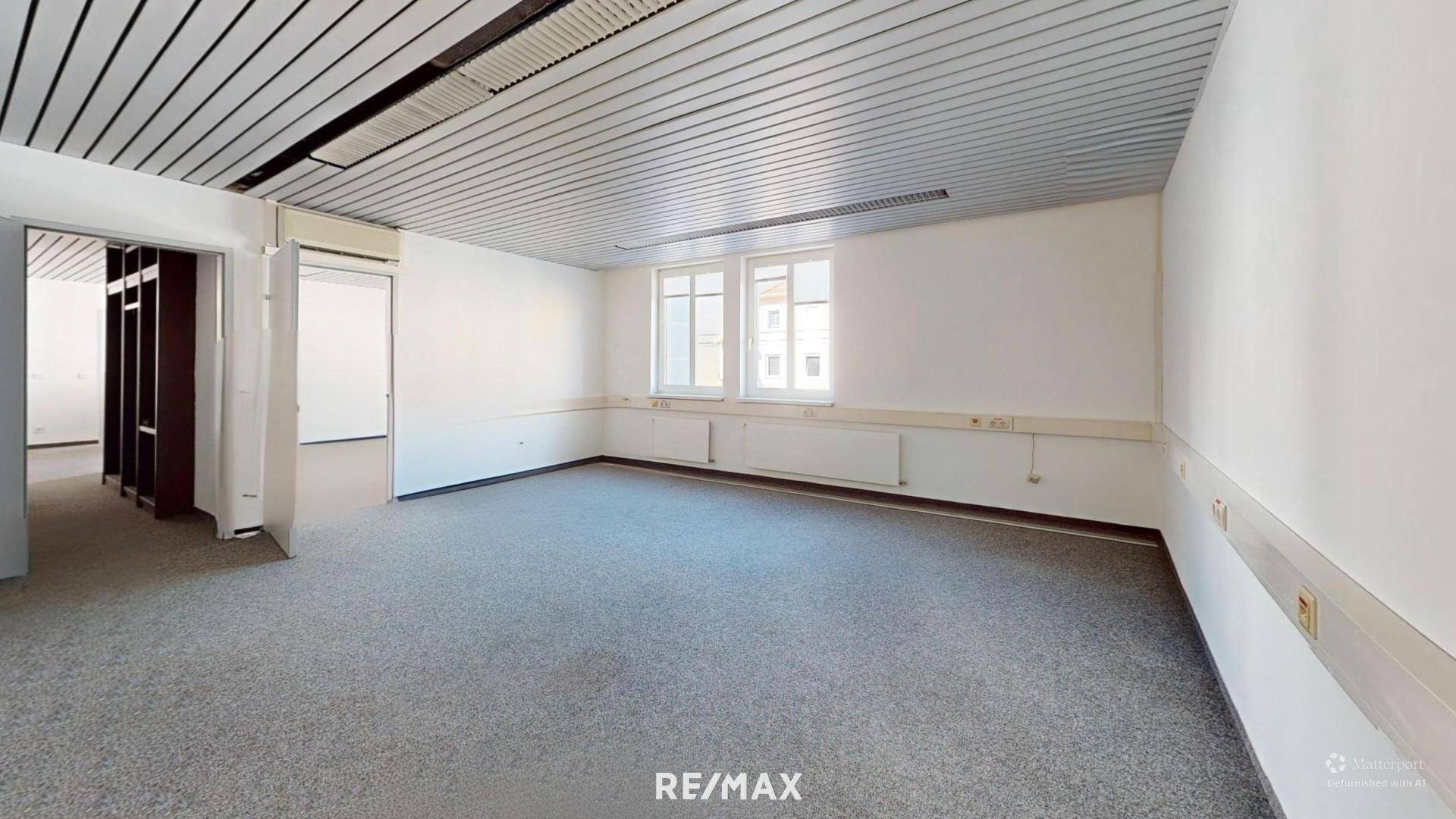 Helles Büro mit Teppichboden, Lamellendecke und Fenstern mit Blick nach draußen.