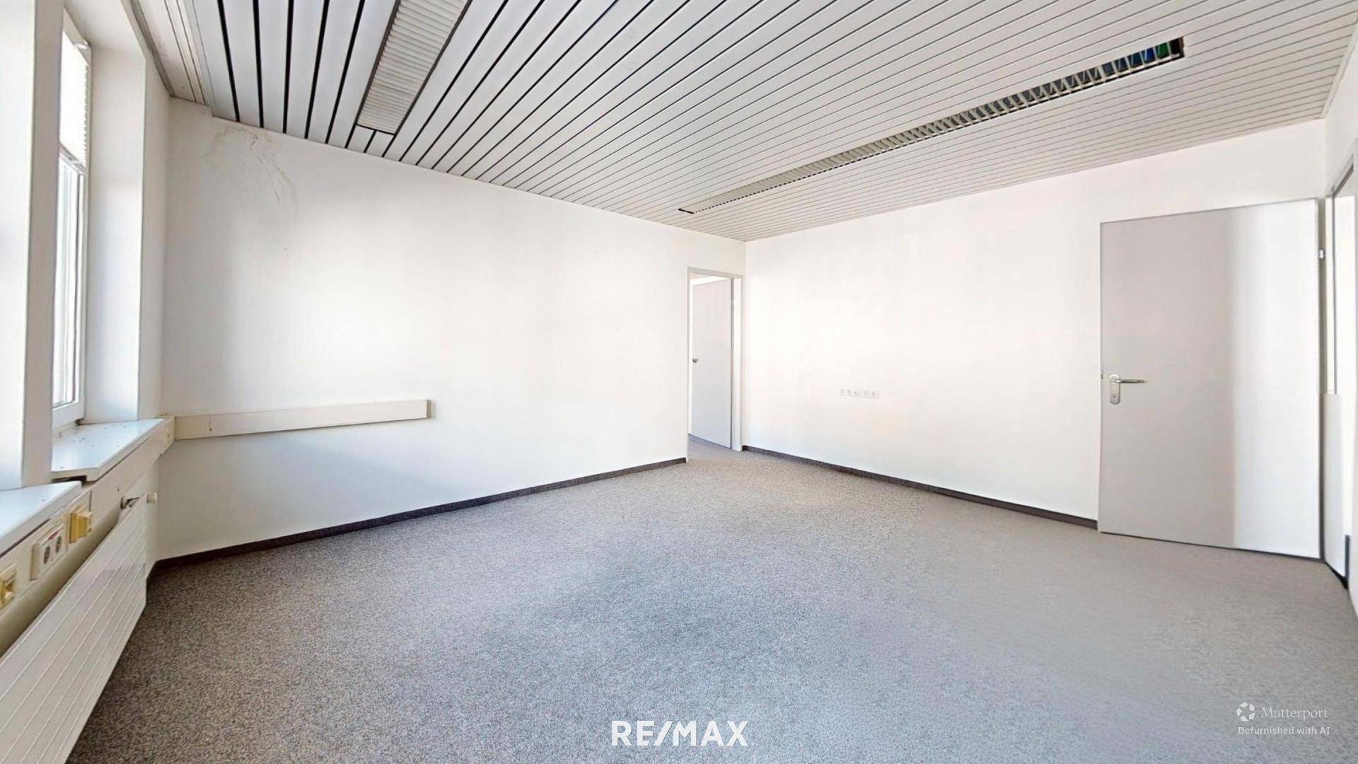 Leeres Büro mit Teppichboden, weißen Wänden und einer Holzleiste entlang der Wand.