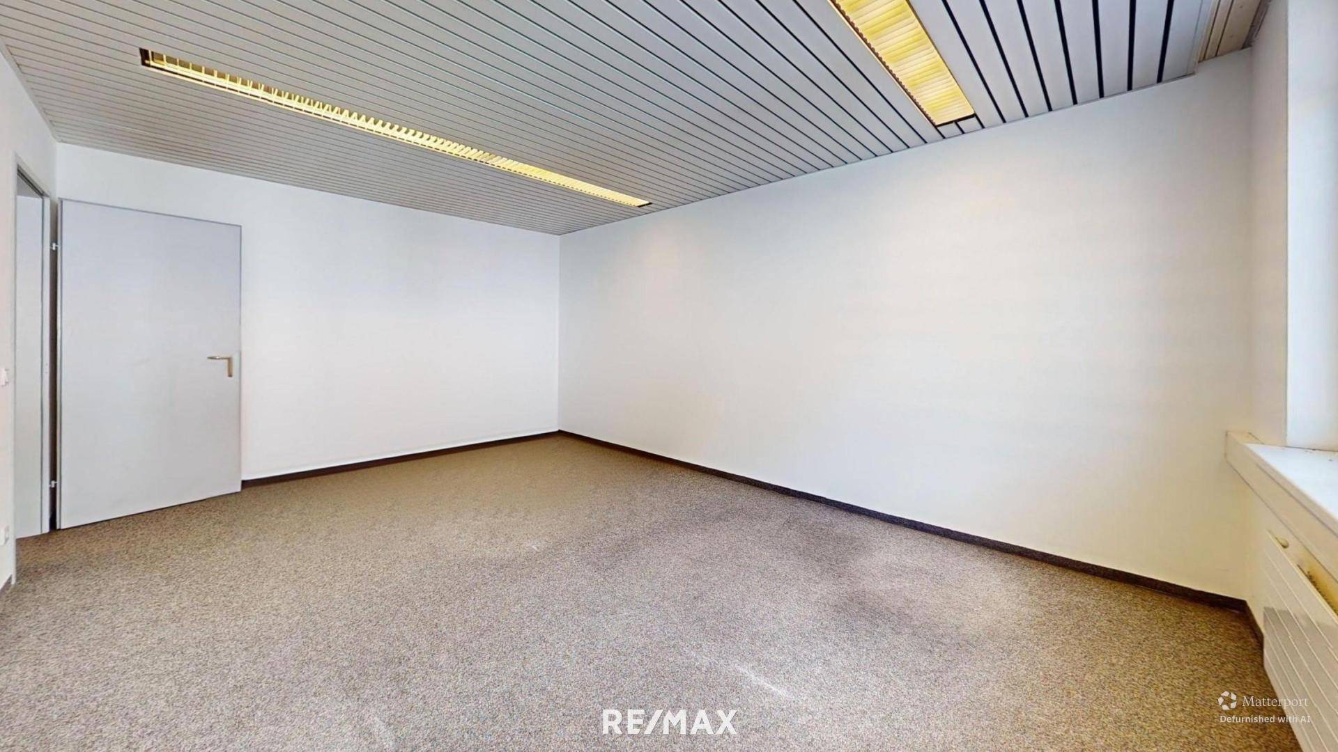 Helles Büro mit Teppichboden, weißen Wänden und einer modernen Deckenbeleuchtung.