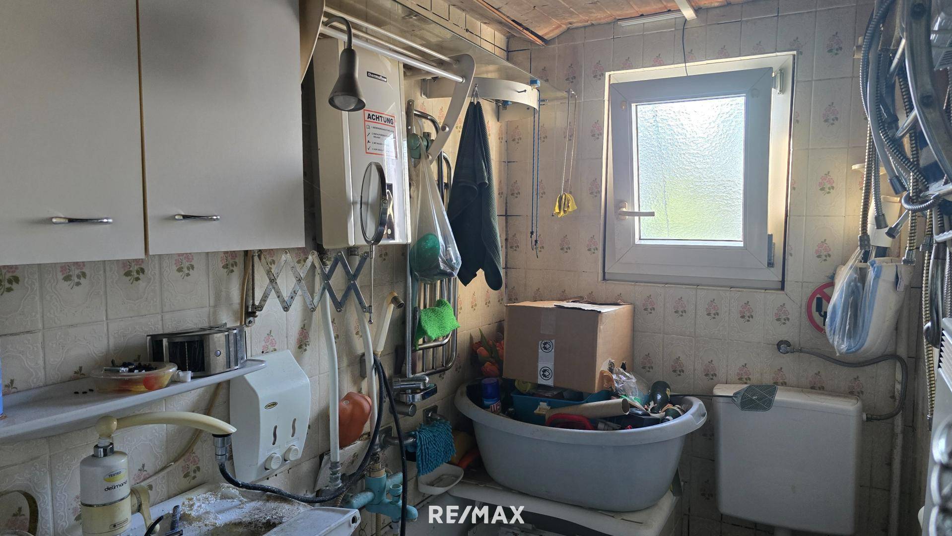 Einfacher Hauswirtschaftsraum mit Waschbecken, Boiler und einem kleinen Fenster.