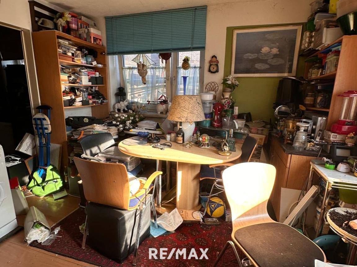 Überladenes Wohnzimmer mit einem runden Tisch, Stühlen und einem Fenster mit Jalousien.