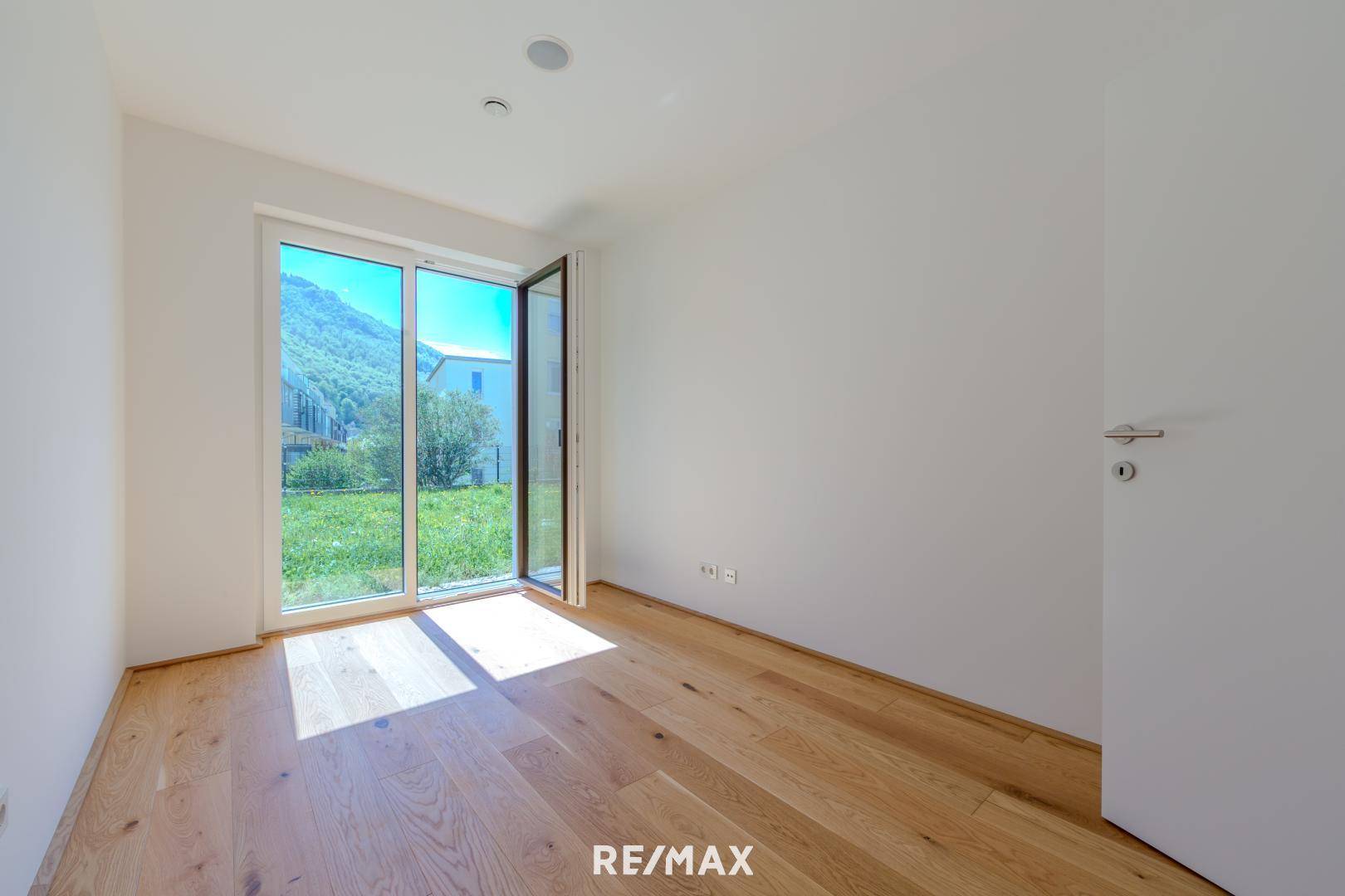 Lichtdurchflutetes Zimmer mit Holzboden und großem Fenster mit Blick auf grüne Natur und Berge.