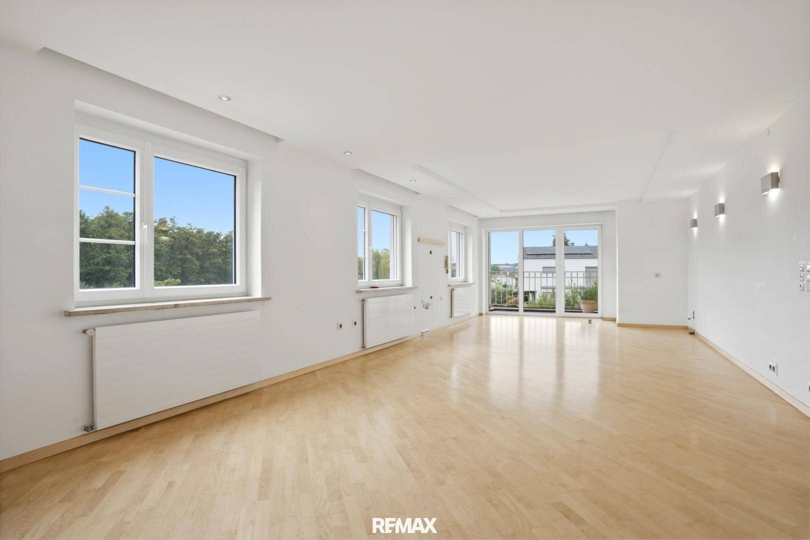 Geräumiges Wohnzimmer mit hellem Parkettboden, vielen Fenstern und Zugang zu einem Balkon.