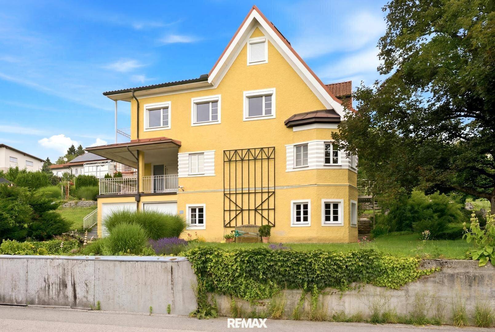Gelbes Wohnhaus mit Balkon und Gartenbereich, von der Straße aus gesehen, umgeben von Grün.
