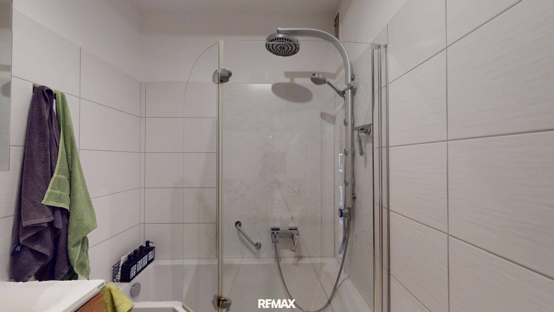 Badezimmer mit Badewanne und integrierter Dusche, ausgestattet mit weißen Wandfliesen und Duschabtrennung.