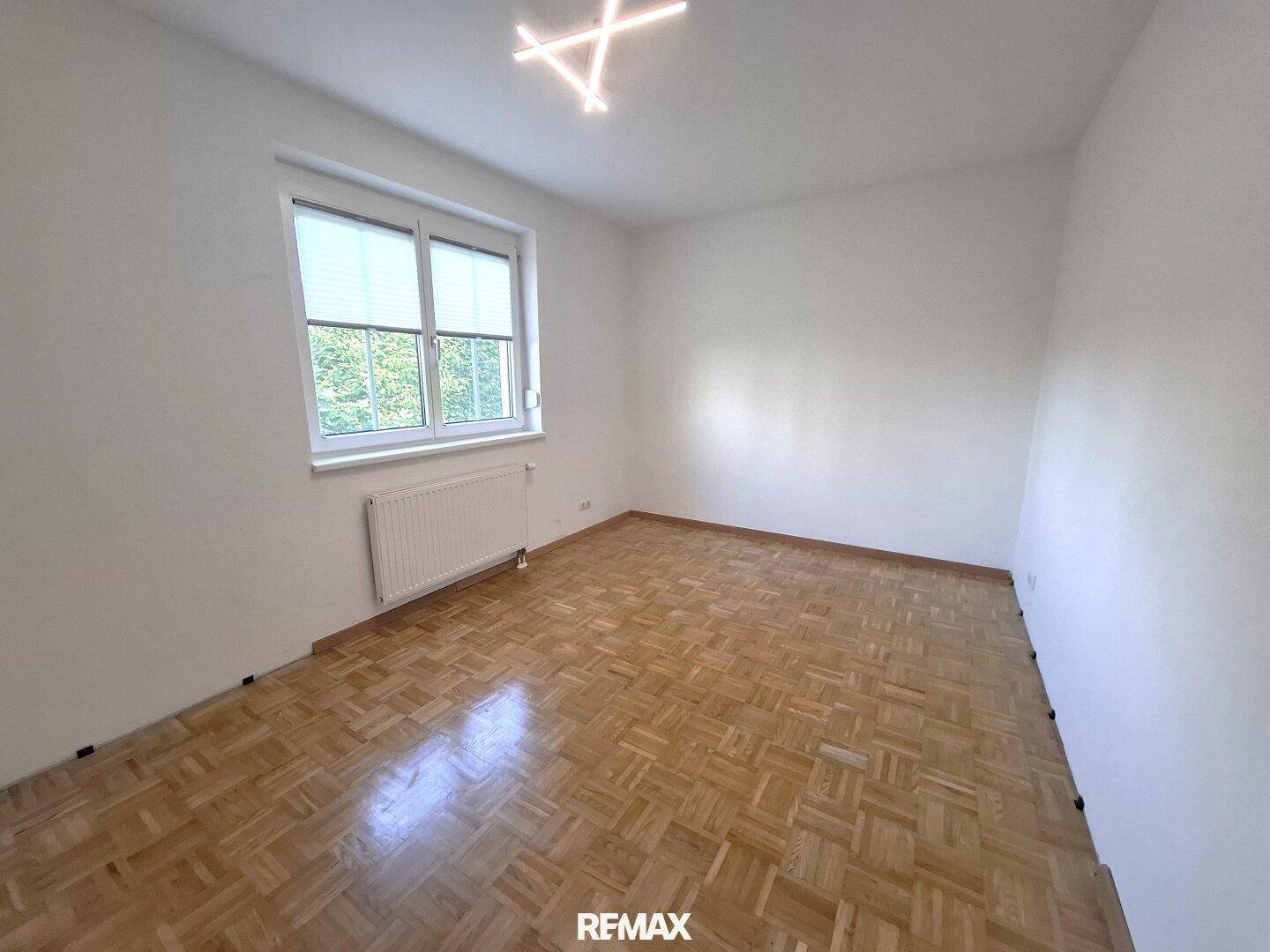 Helles Zimmer mit klassischem Parkettboden, weißen Wänden und Fenster mit Blick ins Grüne.
