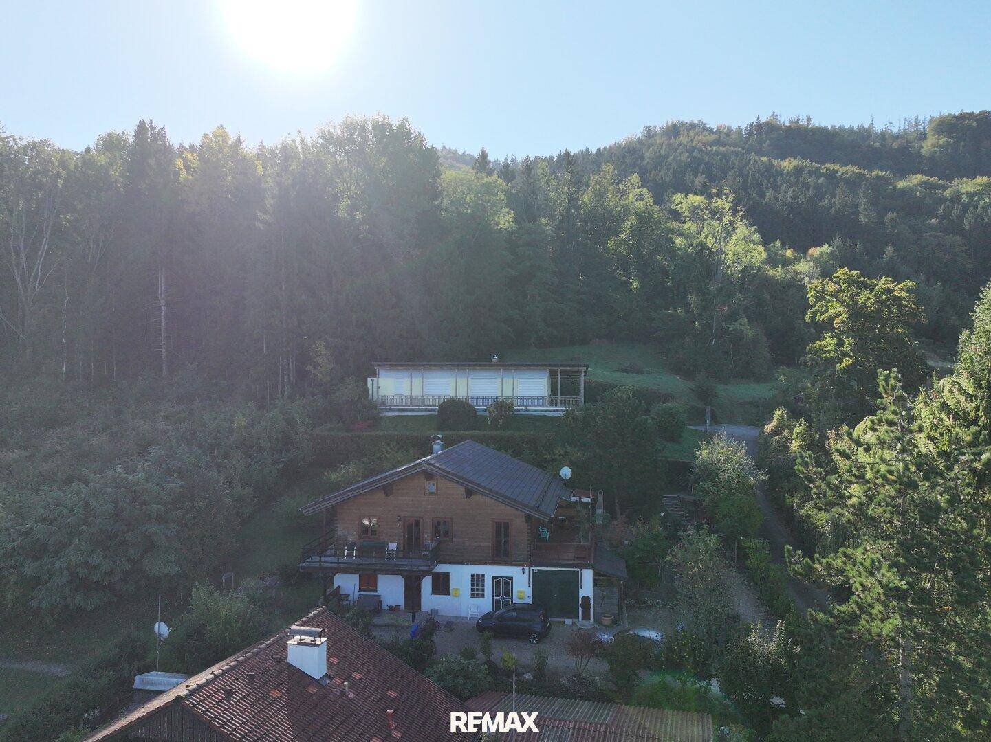 Luftaufnahme des Holzhauses, eingebettet in eine bewaldete Hügellandschaft unter strahlender Sonne.